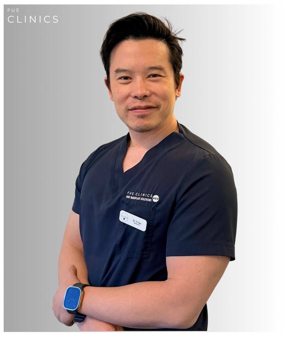 Portrait of a smiling Asian male doctor in navy scrubs with arms crossed. He's wearing a watch against a white background.
