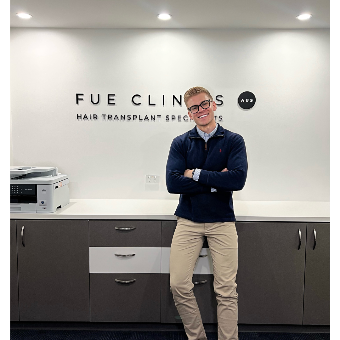 Smiling man in front of FUE Clinics sign. Hair transplant specialist with arms crossed. Clinic interior. FUE hair transplant expert.