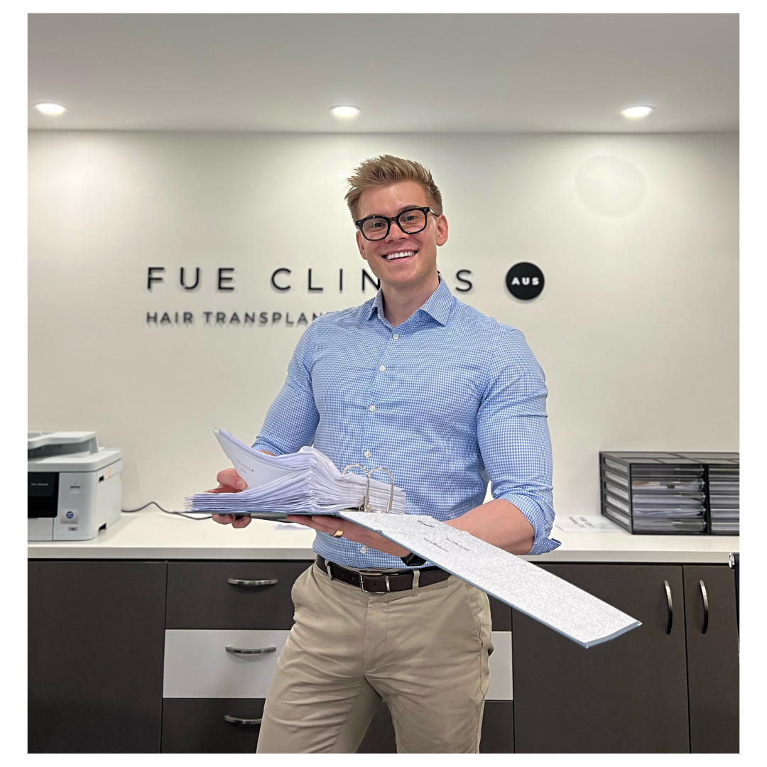 Smiling man holding papers at FUE Clinics, a hair transplant clinic. He's wearing glasses and a blue shirt, standing in front of the clinic's logo.