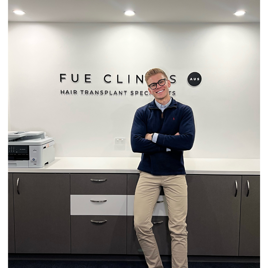 Smiling man in front of FUE Clinics sign. Hair transplant specialist with arms crossed. Clinic interior. FUE hair transplant expert.